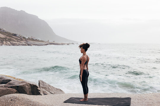 Young female in Mountain Pose outdoors. Woman standing on mat in Tadasana pose by ocean at sunset.