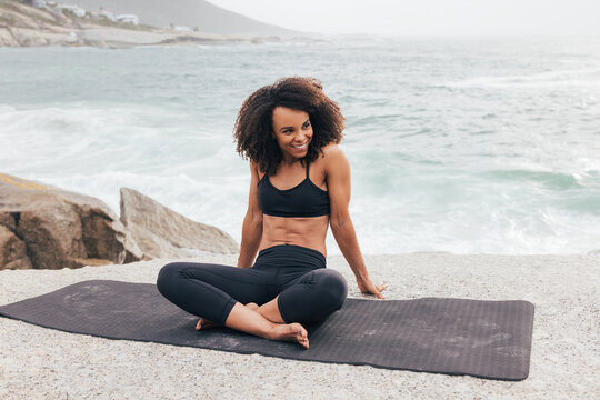 Smiling female relaxing on mat and looking away. Woman with crossed legs in sportswear by ocean.