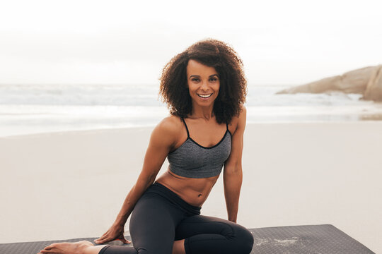 Portrait of a beautiful woman sitting on a mat by ocean. Female with curly hair looking at camera after yoga exercises.