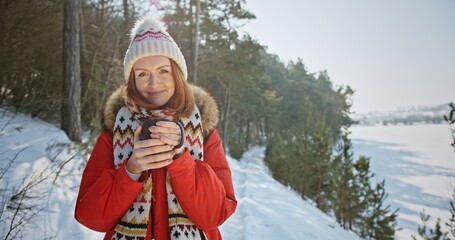 Female traveler enjoying hot drink in winter countryside. Happy young woman in outerwear with scarf and hat warming hands around mug of hot beverage and admiring snowy nature with smile while standing
