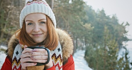 Happy female tourist drinking hot beverage in winter. Cheerful young redhead female in outerwear with knitted hat and scarf sipping hot drink from mug and looking at camera with smile while standing