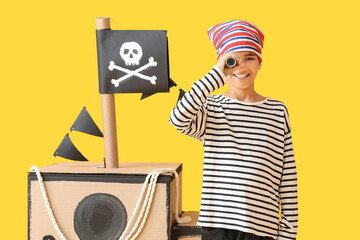 Little boy dressed as pirate with spyglass and cardboard ship on yellow background