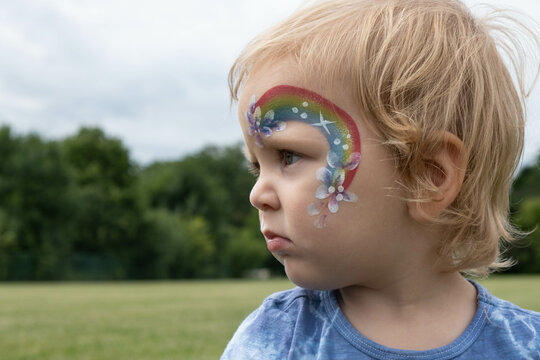 Toddler with rainbow and flower face paint design looks off into the distance; background is lush green park