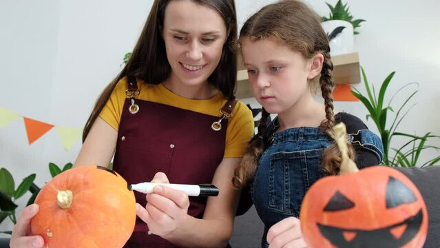 Portrait of cute positive young mother and little daughter sitting at wooden white table and making Halloween decor together at home, painting pumpkins and drawing spooky faces. Holiday concept