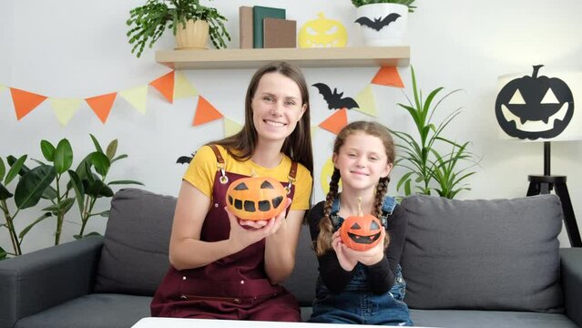Playful smiling mother with funny little daughter girl holding spooky orange Halloween pumpkins in front of face while preparing for Saints Day Party at home, happy mom and kid painting squashes