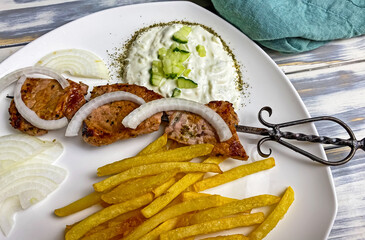 Closeup of white dish with homemade grilled greek pork salmon meat souvlaki, french fries, sweet onions, tzatziki dip, old cast iron skewer, wood table
