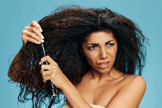 Angry upset curly Latin woman using a hair straightener, posing isolated on blue wall background, look aside. Hair routine concept, haircare, hair ironing, hairdressing. Dry damaged hair problems