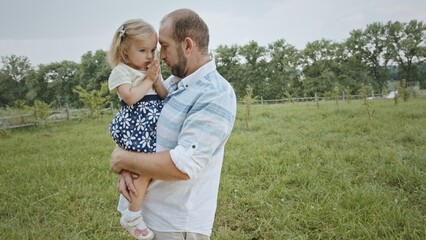Father kissing daughter in field. Adult dad in casual clothes talking and kissing cute daughter while standing in grassy meadow with child in arms on summer weekend day in nature