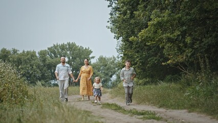Family with dog walking on countryside road together. Adult parents holding hands with son and strolling on countryside road behind daughter and white dog on summer weekend day in countryside