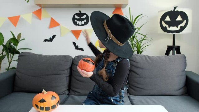 Adorable cute little vlogger in black hat looking at webcam, smiling child girl with pumpkins talking to camera making video call vlog communicating online sit on cozy couch at home. Halloween concept