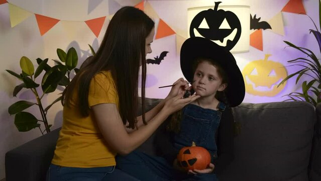Loving young mom painting face cute little daughter child  for Halloween celebration, family sitting together on cozy couch in dark living room decorated spooky paper black spooky pumpkins and bats