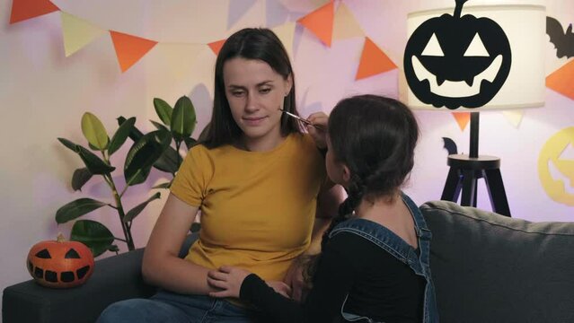 Fun cute little daughter child painting face cute young mom for Halloween celebration, family sitting together on comfy sofa in dark living room decorated spooky paper black spooky pumpkins and bats