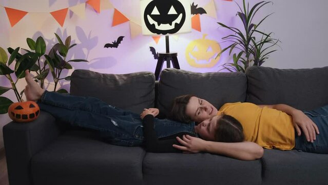 Relaxed young mom and cute kid daughter telling story during Halloween celebration, caring mother and little child girl lying on cozy sofa in living room decorated paper spooky pumpkin and small bats