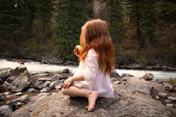 a little girl with long red hair in a white dress is sitting near the river and eating an apple