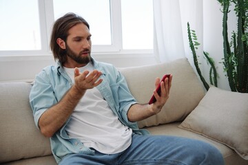 Obraz premium A young man looks angrily and sadly at the phone screen at home sitting on the couch. Bad news and bad luck at work