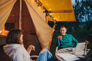 Happy family couple freelancers have a rest after working laptop on cozy glamping tent in summer evening. Luxury camping tent for outdoor holiday and vacation. Lifestyle concept