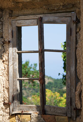 Blick auf durch ein altes Holzfenster, welches von der Sonne angestrahlt wird, grüne Bäume und blauer Himmel dahinter, vertikal 