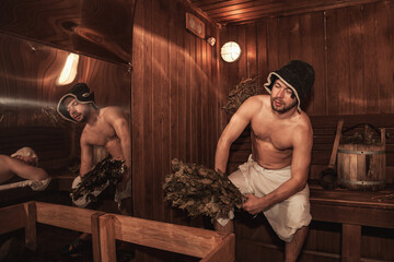 Two men relaxing and sweating in wooden sauna with hot steam. Russian bathhouse. Guys with bath...