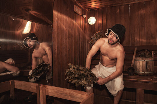 Two men relaxing and sweating in wooden sauna with hot steam. Russian bathhouse. Guys with bath besoms resting on bench in spa complex. Wellness, self care, healthy concept. Copy text space