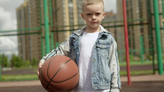 Portrait of boy is standing on basketball court holding ball in city district.