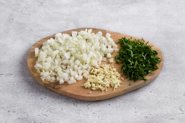 Wooden board with chopped vegetables: onion, parsley and garlic on gray textured background, top view. Cooking homemade food