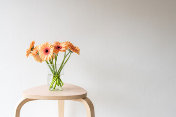 Closeup of orange gerbera daisies in glass vase on wooden stool against beige wall with copy space
