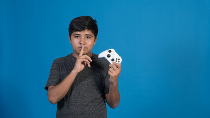 Retrato de estudio de un niño jugando a videojuegos haciendo una señal de silencio con joystick. Fondo Azul. - obrazy, fototapety, plakaty