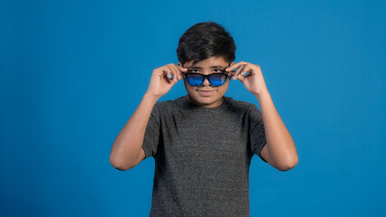 Retrato de estudio de un niño sonriente que se quita las gafas de sol sobre fondo azul.