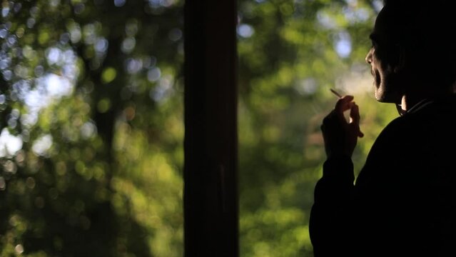 Guy smokes by window. Person smokes cigarette and looks out window. Smoking is harmful to health. Silhouette of man in early morning. Bad habit.