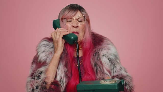 Crazy tourist hacker elderly old woman talking on wired vintage telephone of 80s, fooling, making silly faces. Senior stylish grandmother granny hotline agent isolated alone on pink studio background