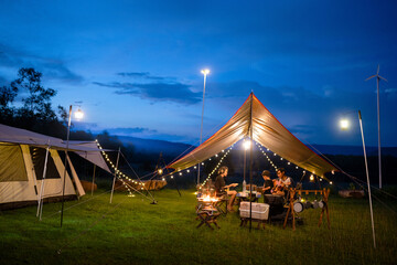 Night camping scenes, long exposures in moving parts are blurry..Night camping with tent accommodation and relaxation seats, with an Asian family having dinner, playing guitar and having fun.