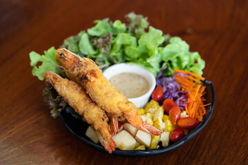 fried or battered shrimp salad with apple corn carrot tomato with green vegetables and salad dressing on black dish and wooden table for diet food or healthy cuisine at restaurant on top view