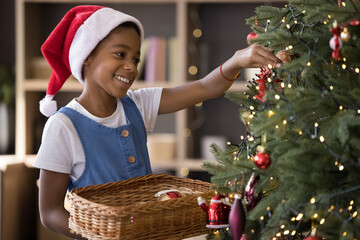 Happy cute African American kid in red Santa cap hanging up ornament baubles on Christmas fir tree...