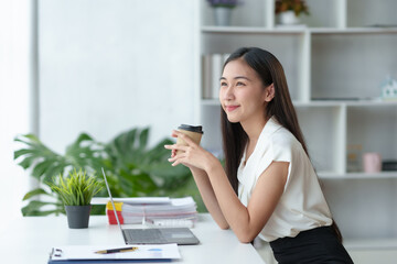 At the office, a young Asian businesswoman sit and relaxe with a cup of coffee and a bright smile on her laptop.