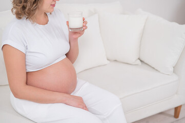 Obraz na płótnie Canvas A pregnant woman sits on a white sofa and drinks milk.