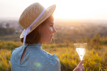 A woman in a blue dress and straw hat with short hair sitting on a white blanket holding glass and drinking white wine. Concept of having picnic in a city park during summer holidays or weekends. 