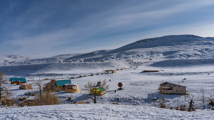In the snow-covered valley, you can see the wooden houses of the campsite and spherical glamping tents. A mountain range against a background of blue sky and cirrus clouds. Altai. Kyzyl Chin