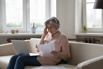 Frustrated annoyed senior woman reading paper bill, invoice, financial document at home, holding...