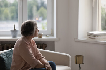 Thoughtful lonely retired woman sitting on couch at home, looking at window away in deep thoughts,...