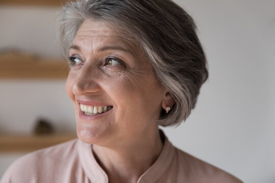 Happy dreamy retired grey haired woman looking away, with toothy smile, thinking, feeling joy. Face of cheerful thoughtful elder 60s lady close up portrait. Elderly age, retirement