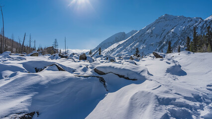 Fototapeta na wymiar Boulders piled up in the valley are covered with a thick layer of pure white snow. Mountains against a clear blue sky. The sun is shining in the firmament. Altai