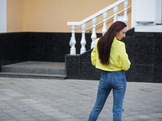 A stylish slender sexy woman stands near the stairs and stone railings on the city street. She is wearing blue jeans and a short yellow leather jacket.