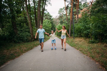 A happy young family, dad, mom and son are skating hand in hand with their parents, on roller skates, along a path in the park. The whole family is dressed in shorts and T-shirts.