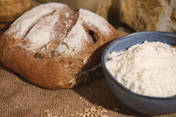 Selective focus on fresh baked homemade traditional sourdough bread, and a blue ceramic bowl with white flour on burlap