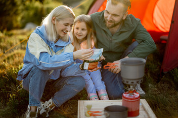 Young couple with little girl have picnic, cooking dried-food for hiking in package at campsite. Concept of sublimation food for travel and family bonding on travel