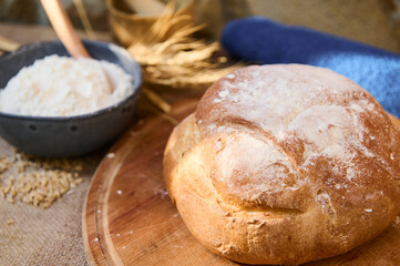 A loaf of fresh baked homemade whole grain sourdough bread, on a wooden board, bowl with flour and spikelets of wheat.