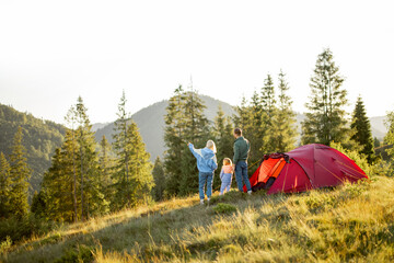 Family with a kid enjoy great view on mountains while stand together at campsite near the tent. Family trip on nature concept