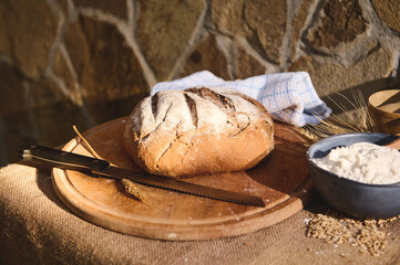 Still life. Multigrain loaf of bread and kitchen knife on a wooden board, next to a bowl with white flour and wheat ears
