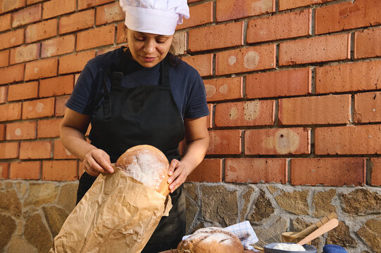 Beautiful woman baker, shop assistant in artisan bakery shop, packing fresh sourdough wheaten bread in eco bag, for sale