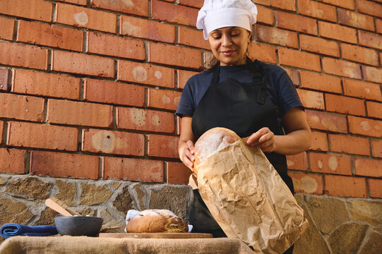 Charming woman baker, shop assistant in artisan bakery shop, packing fresh sourdough wheaten bread in eco bag, for sale
